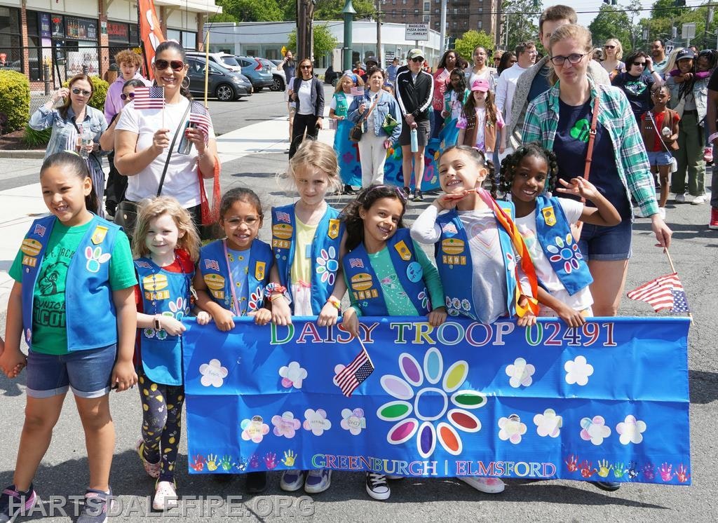 A group of young girls in blue uniforms holding a Daisy Troop banner during a parade, with adults and onlookers in the background.