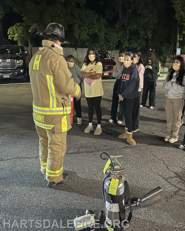 A firefighter is teaching a group of masked individuals about fire safety, with a fire extinguisher displayed in front.