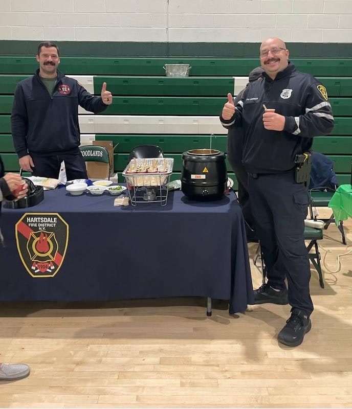 Two smiling men, one in a fire department uniform and the other in police attire, stand by a food table.