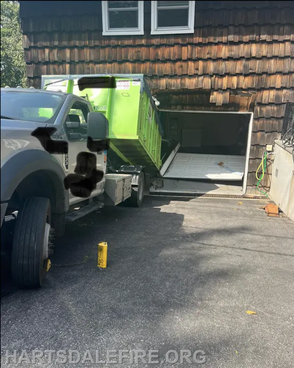 A truck with a green dumpster is partially inside a garage, causing the garage door to be raised and damaged.