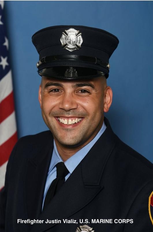 A smiling firefighter in uniform, wearing a hat with an emblem, posed in front of a blue backdrop and an American flag.