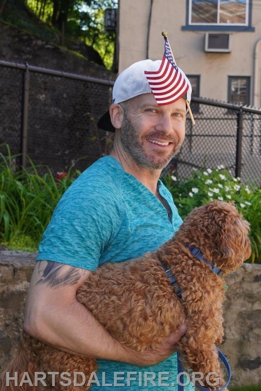 A smiling man in a blue shirt holds a dog while wearing a small American flag hat. Greenery and a building are in the background.