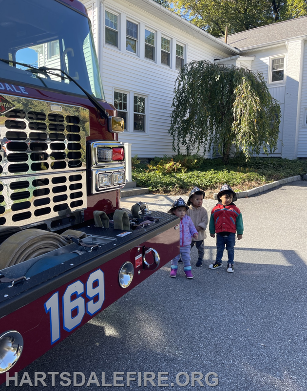 Two young children wearing hats stand next to a fire truck in front of a house. It's a sunny day.
