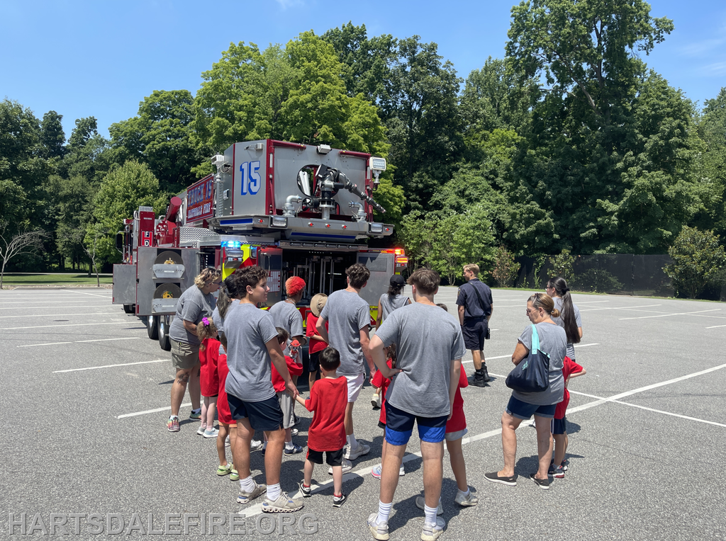 A group of children and adults gathers around a fire truck on a sunny day, likely for a community event or demonstration.
