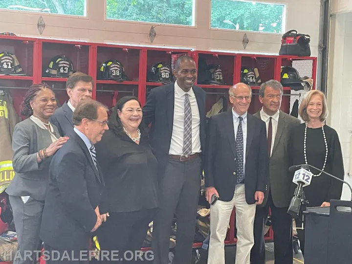 Group of people posing in front of firefighter gear, some smiling, with a microphone stand nearby.