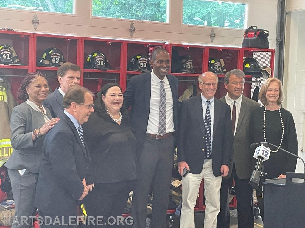 Group of people posing in front of firefighter gear, some smiling, with a microphone stand nearby.