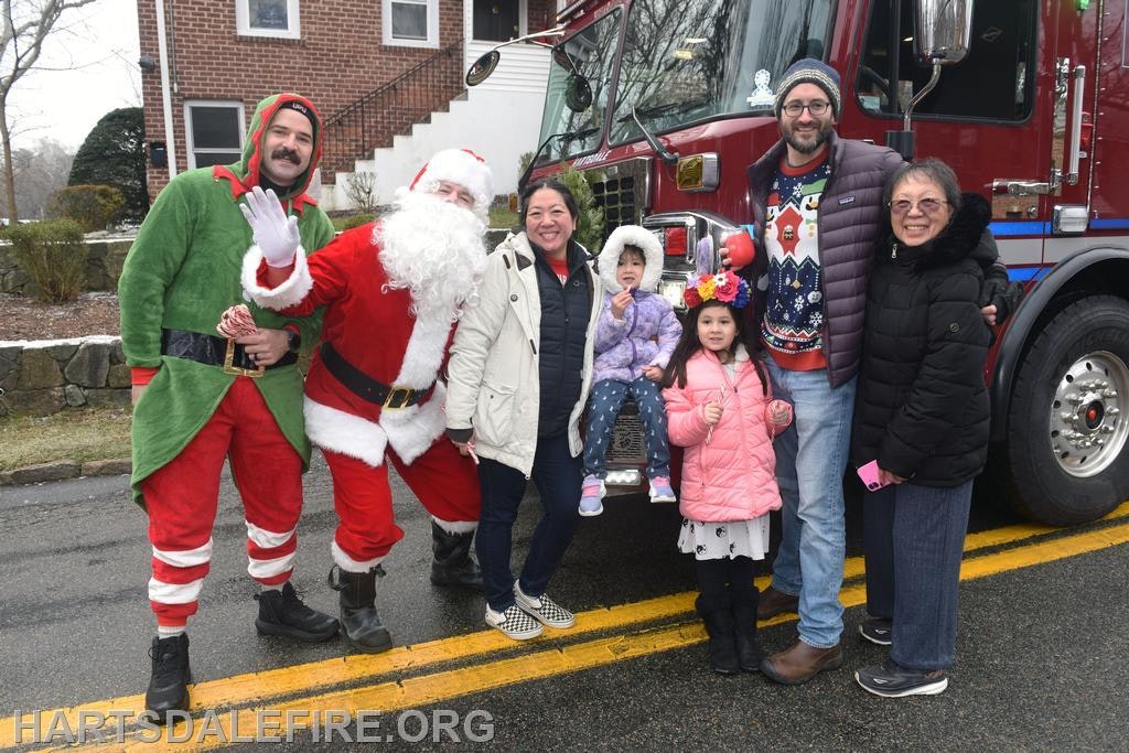 A festive group poses with Santa, an elf, and a fire truck, celebrating the holiday spirit with children and family.