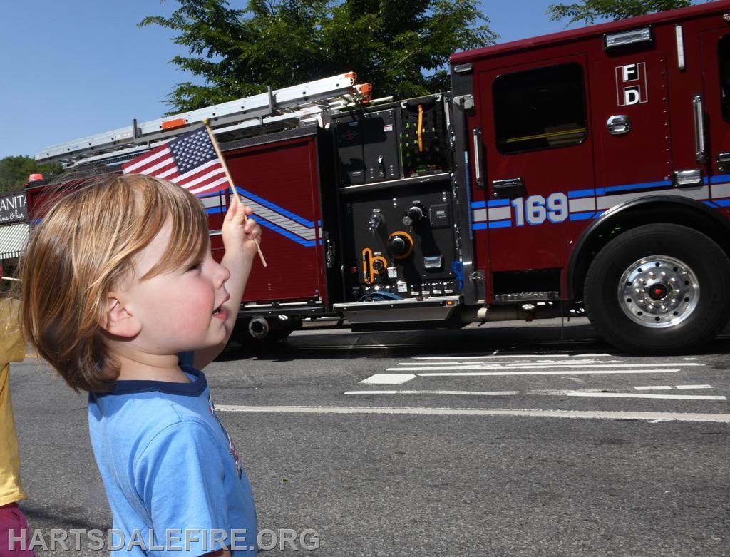 A child waves an American flag at a passing red fire truck labeled “169”.