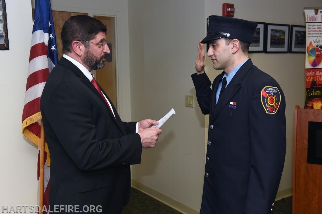 A person in a suit reads to a firefighter in uniform, who's raising their right hand, in front of a flag.