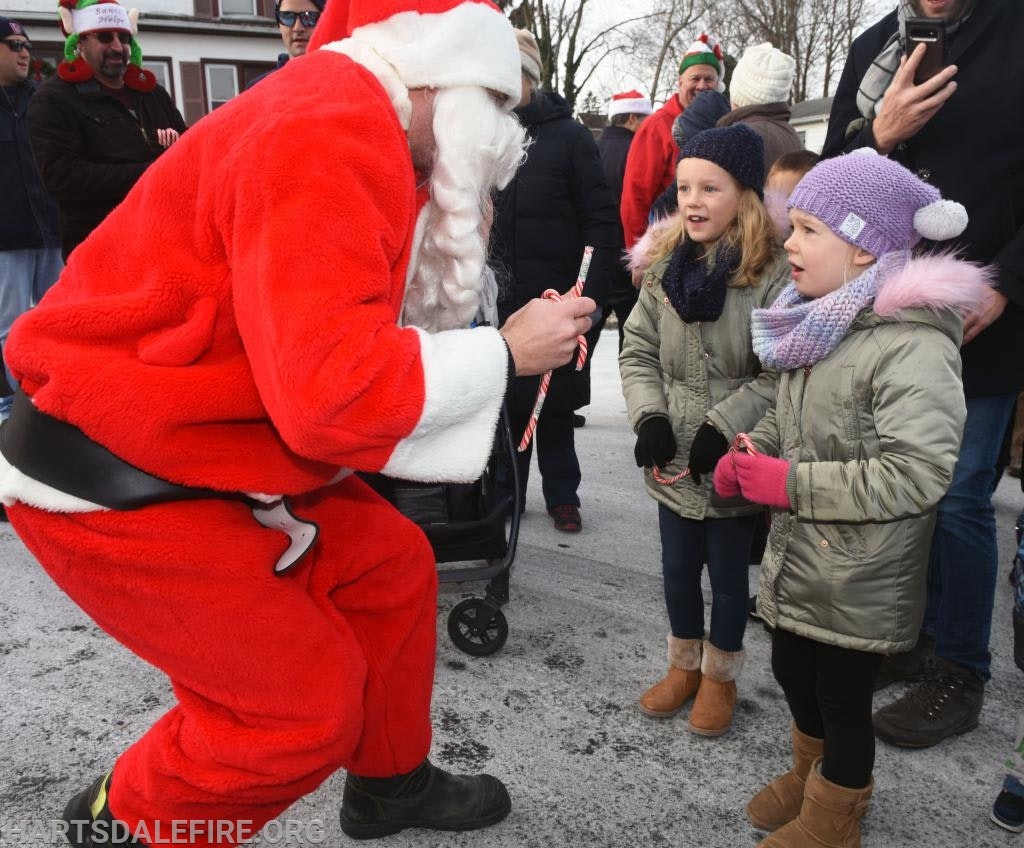 Santa gives candy canes to two children in winter clothing at an outdoor event.