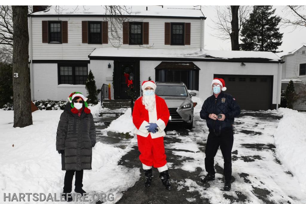 Three people in festive outfits and masks stand in a snowy driveway in front of a house.