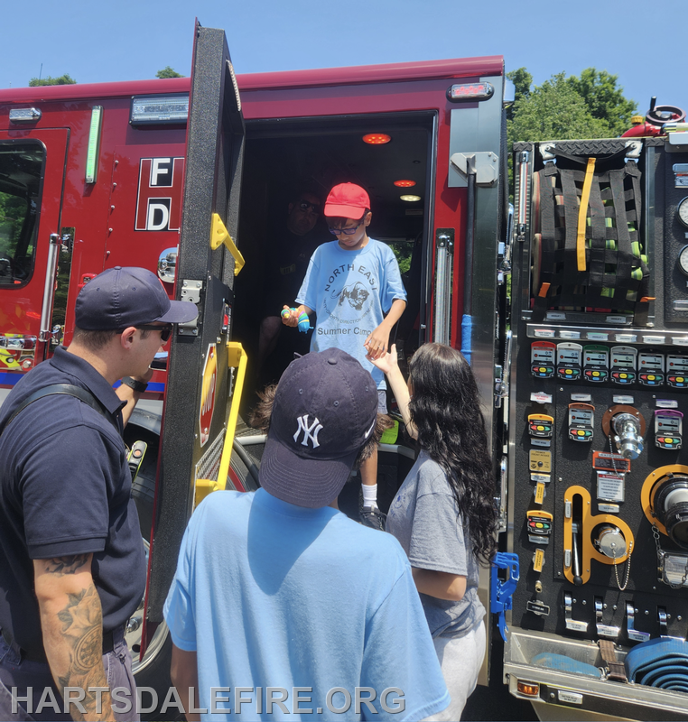 A child in a red cap exits a fire truck, interacting with adults nearby on a sunny day, engaging in a community event.