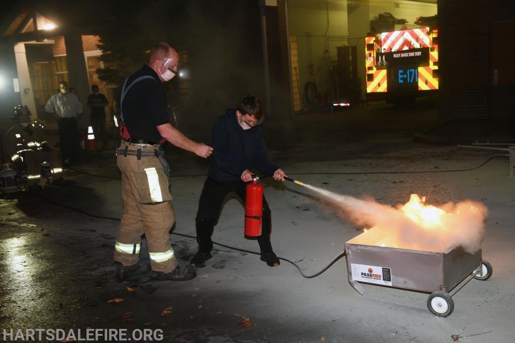 A person uses a fire extinguisher on a controlled fire, assisted by a firefighter in a training setting.