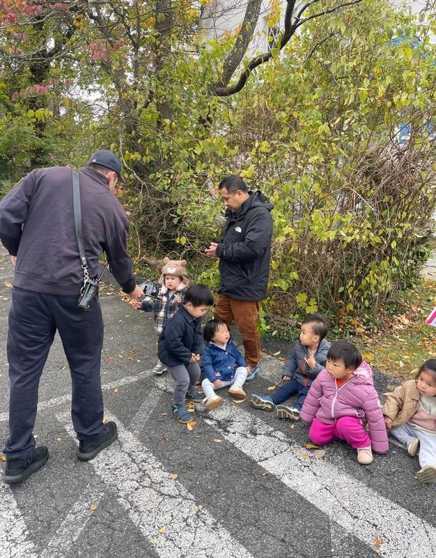 A group of young children sits on the ground as an adult hands something to one child, with another adult observing nearby.