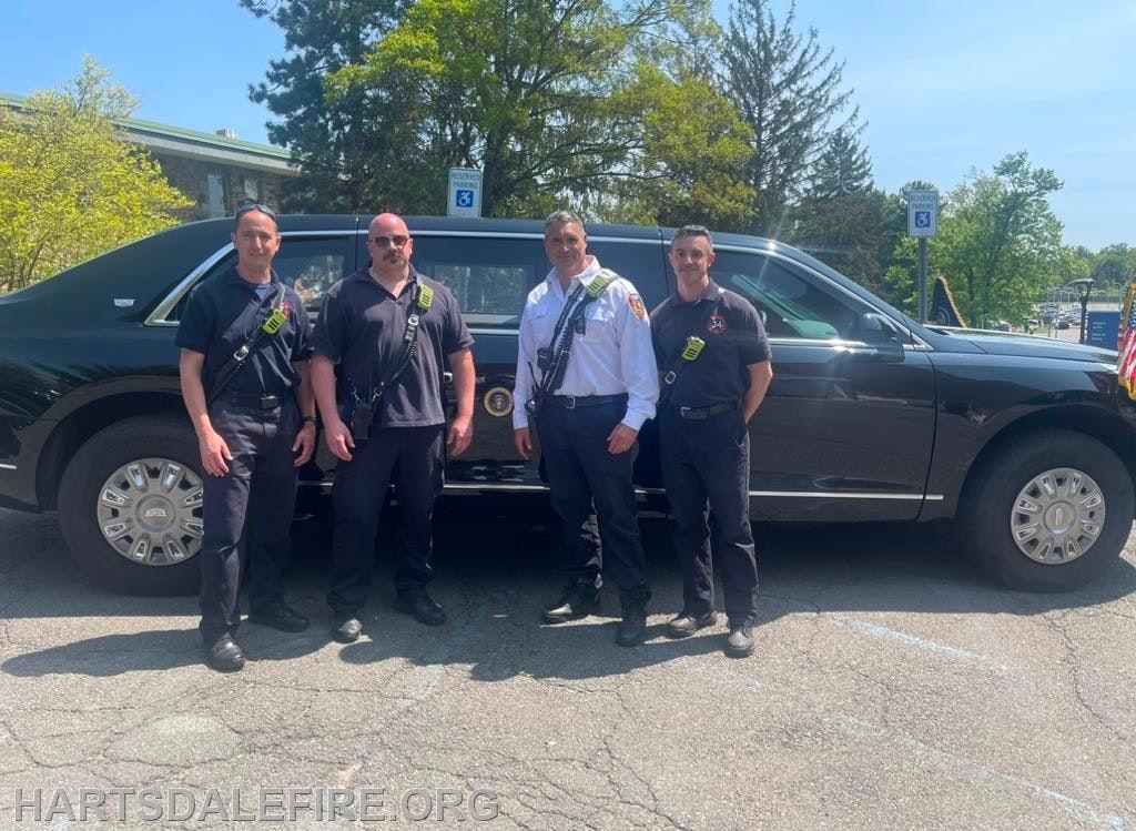 Four firefighters stand in front of a black car with emergency parking signs visible.