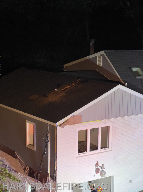 The image shows a house with significant damage to the roof, likely from an incident, illuminated at night.
