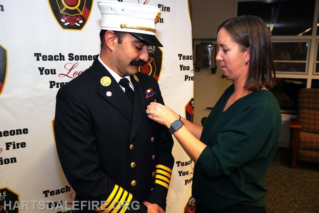 A woman pins a badge on a man in a fire captain's uniform at a ceremony, with a supportive backdrop.