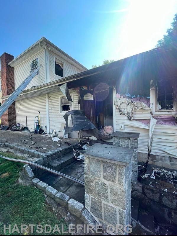 House with visible fire damage, charred walls, debris on the ground, and a ladder positioned against the upper window.