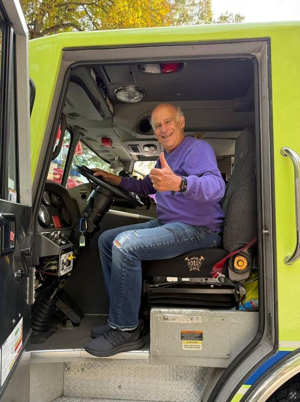 A smiling man in a purple sweater gives a thumbs-up while seated in the driver's seat of a green vehicle, likely an emergency vehicle.