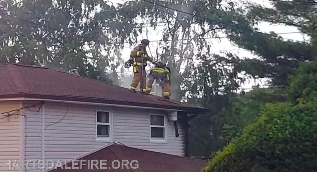 Firefighters on a house roof with equipment, managing smoke.