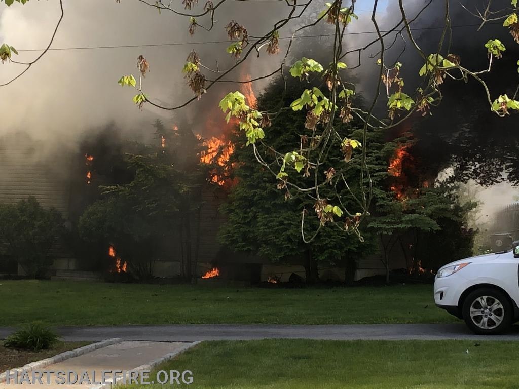 A house is on fire with visible flames and smoke; a white SUV is parked on the side.