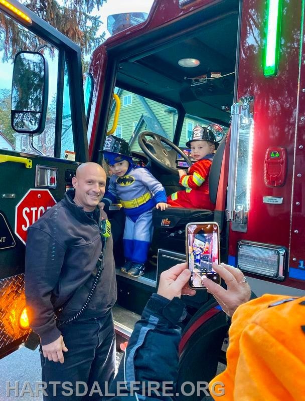 A fire truck scene featuring a smiling firefighter and two kids in costumes, one as Batman, excitedly posing for a photo.