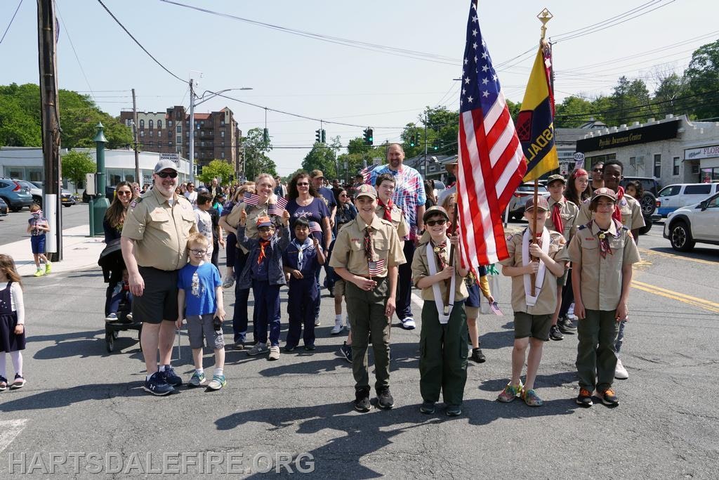 A group of scouts and adults in a parade, holding American flags on a sunny street.