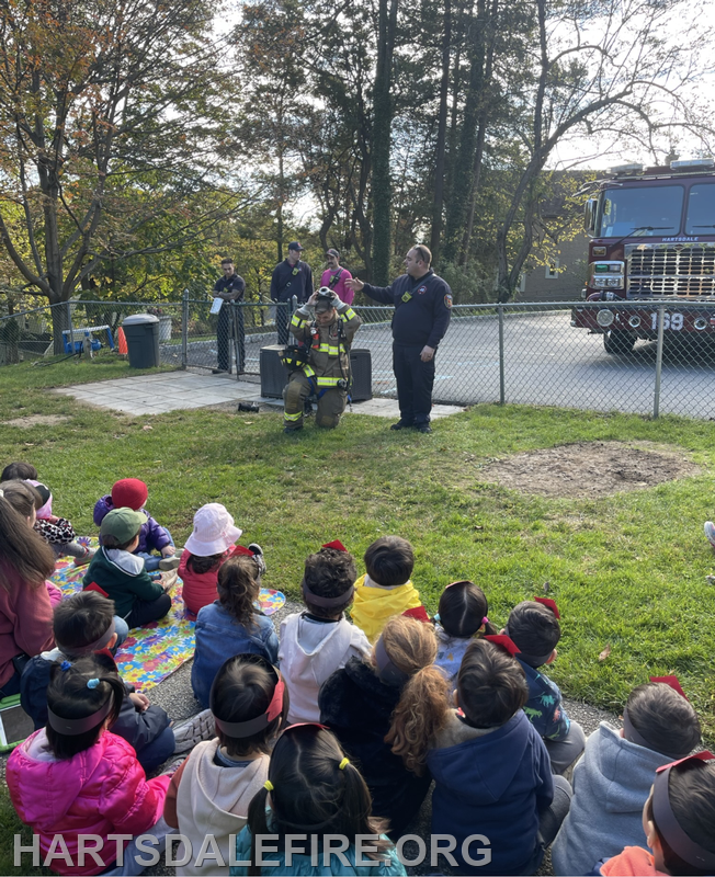 A firefighter demonstrates safety while children watch attentively during a community event. A fire truck is in the background.