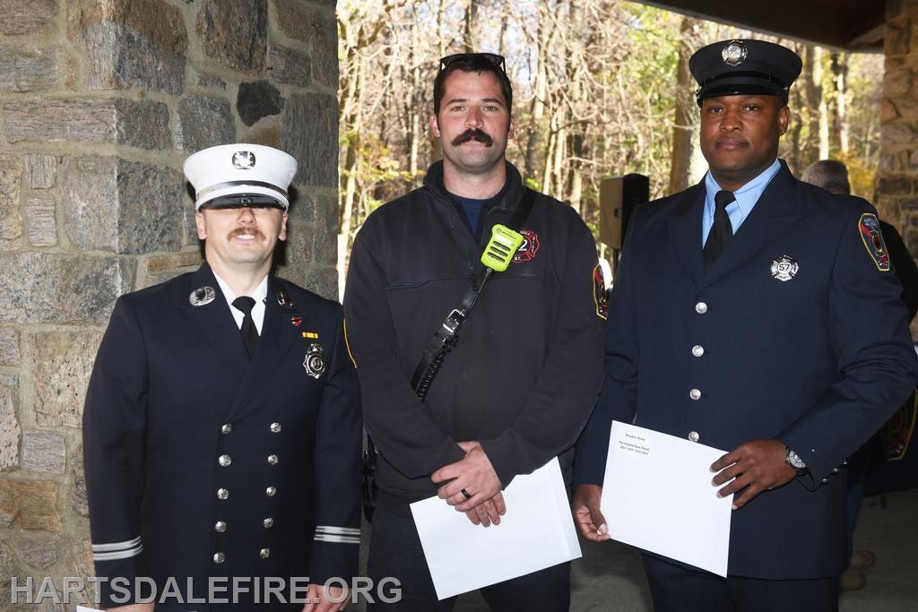 Three men in formal attire, including firefighters, stand together outdoors, smiling. They hold papers and appear to be at an event.