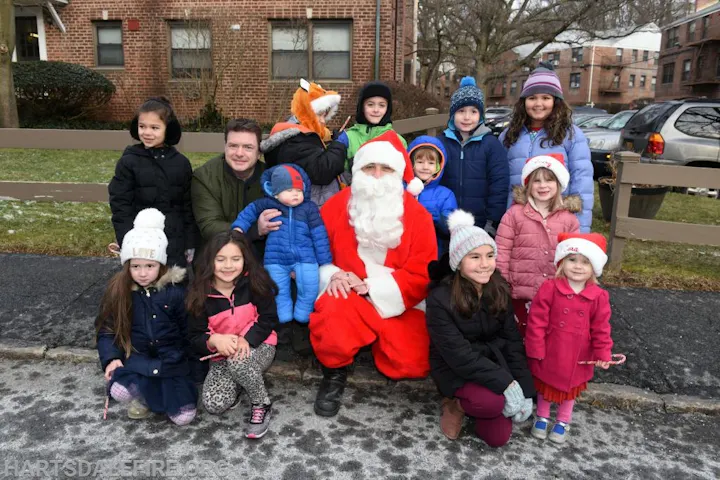 A group of children in winter clothing pose outdoors with Santa Claus in a festive setting.