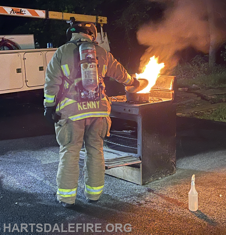 A firefighter in gear is extinguishing a fire on a stovetop outdoors at night. A spray bottle is nearby.