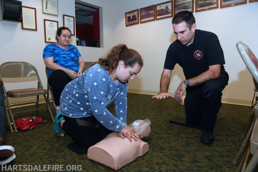 A person demonstrates CPR on a mannequin while another observes and an instructor guides them.