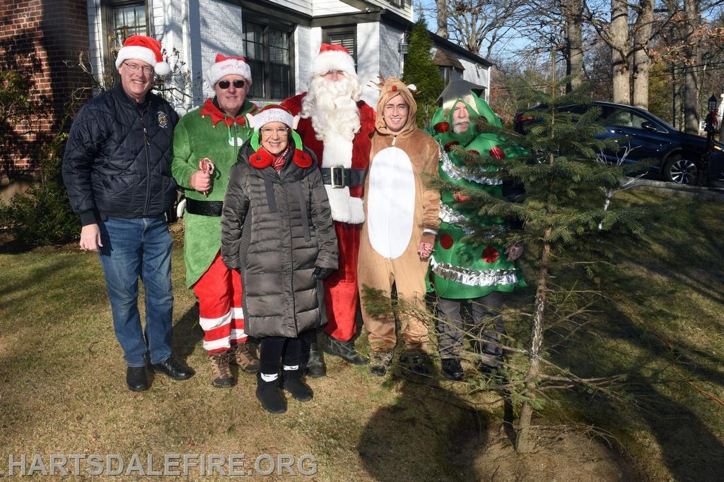 A festive group of people in holiday costumes, including Santa, elves, and a reindeer, gathered outdoors near a tree.