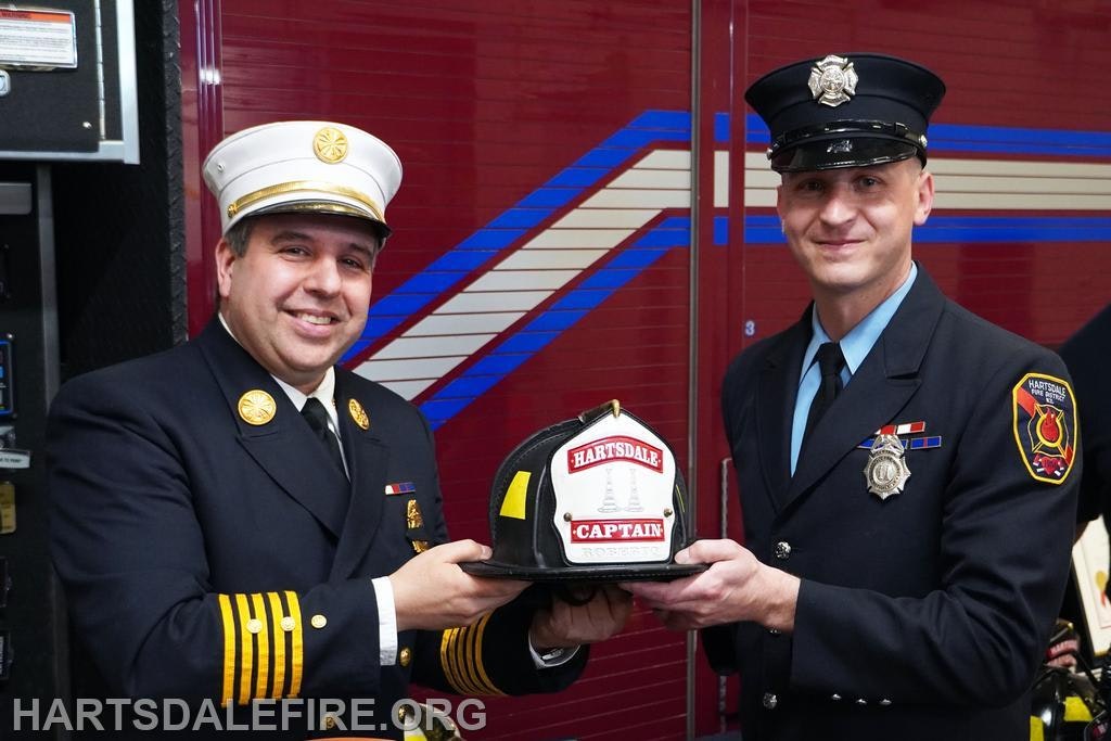 Two firefighters in uniform holding a captain's helmet, standing in front of a fire truck.