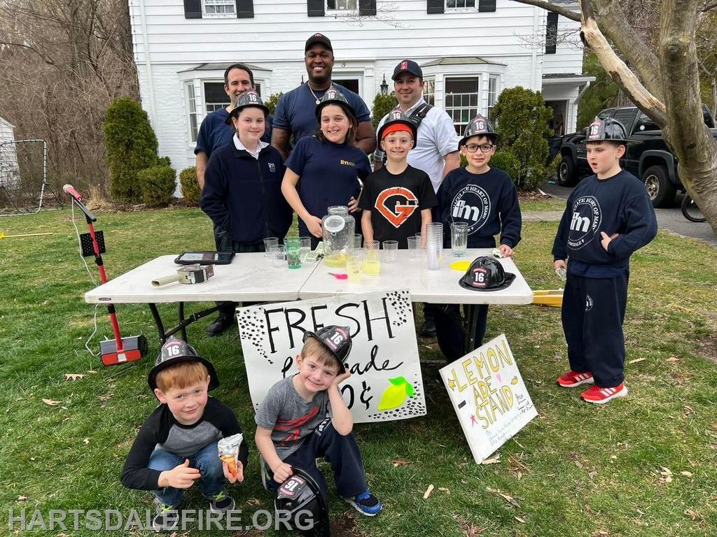 A group of kids and adults run a lemonade stand, promoting "Fresh Lemonade" in a friendly neighborhood setting.