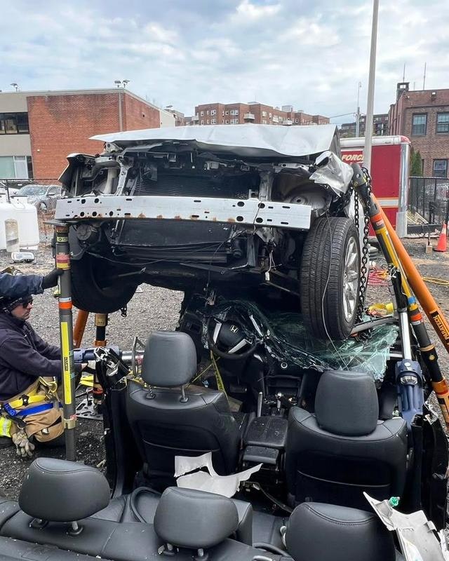 A car is flipped upside down, resting on another vehicle, with emergency responders working nearby.