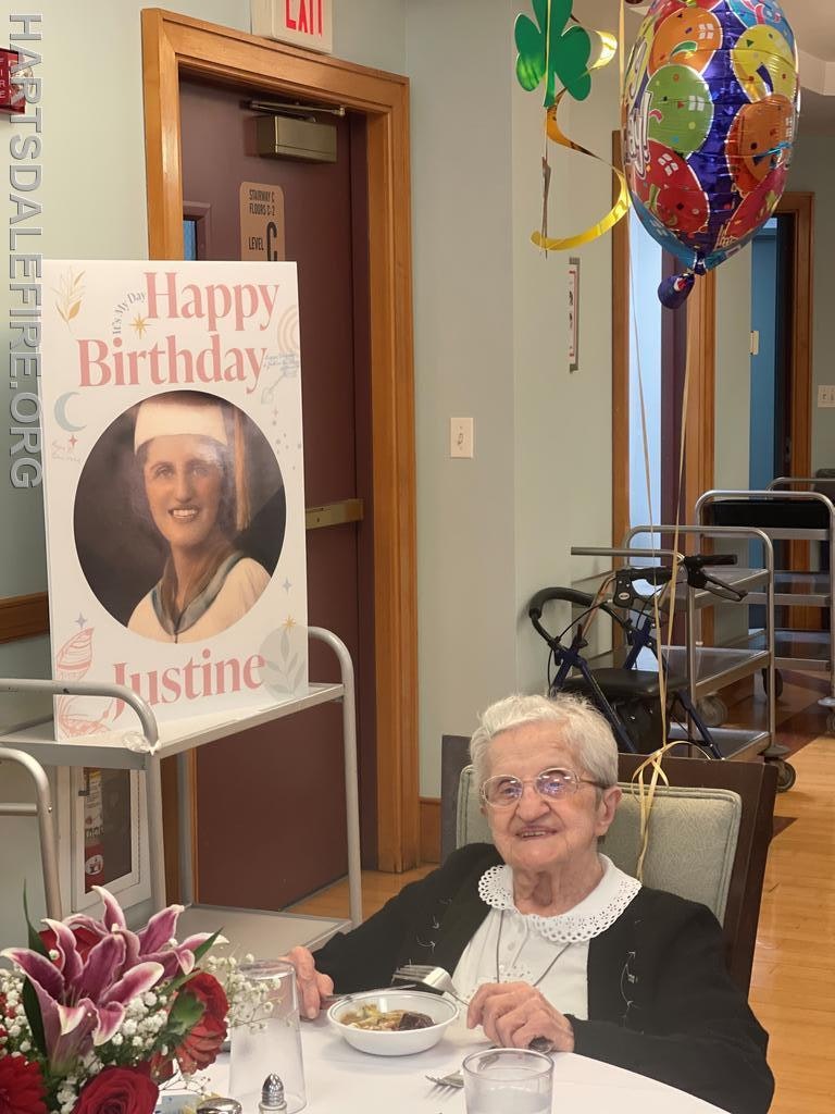 A elderly woman celebrates her birthday with a meal, surrounded by festive decorations and a birthday sign.