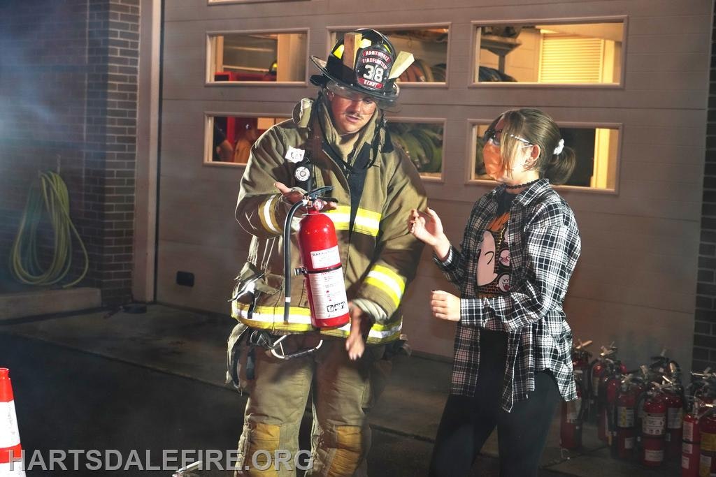 Firefighter demonstrating fire extinguisher use to a person, both wearing safety gear, in front of a building.