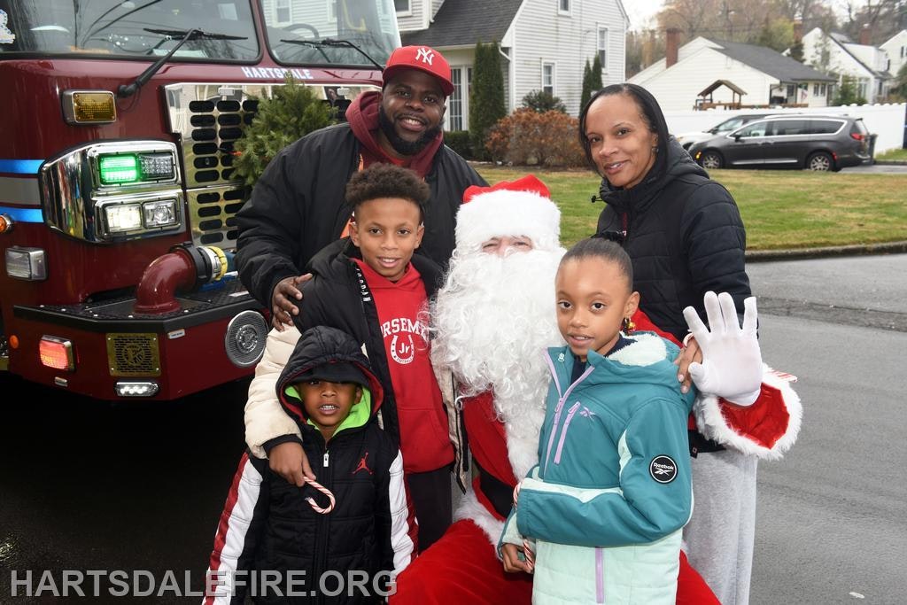 A family poses with Santa in front of a fire truck during a festive community event. Kids are smiling and holding candy canes.
