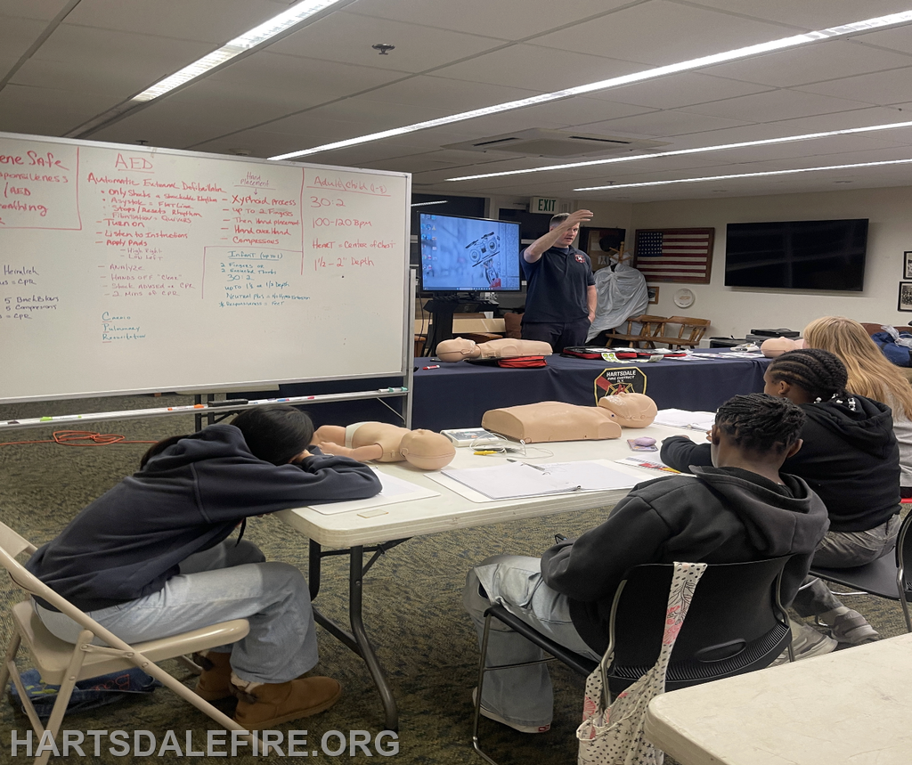 A group learning CPR, with mannequins, a whiteboard with notes, and a speaker demonstrating techniques.