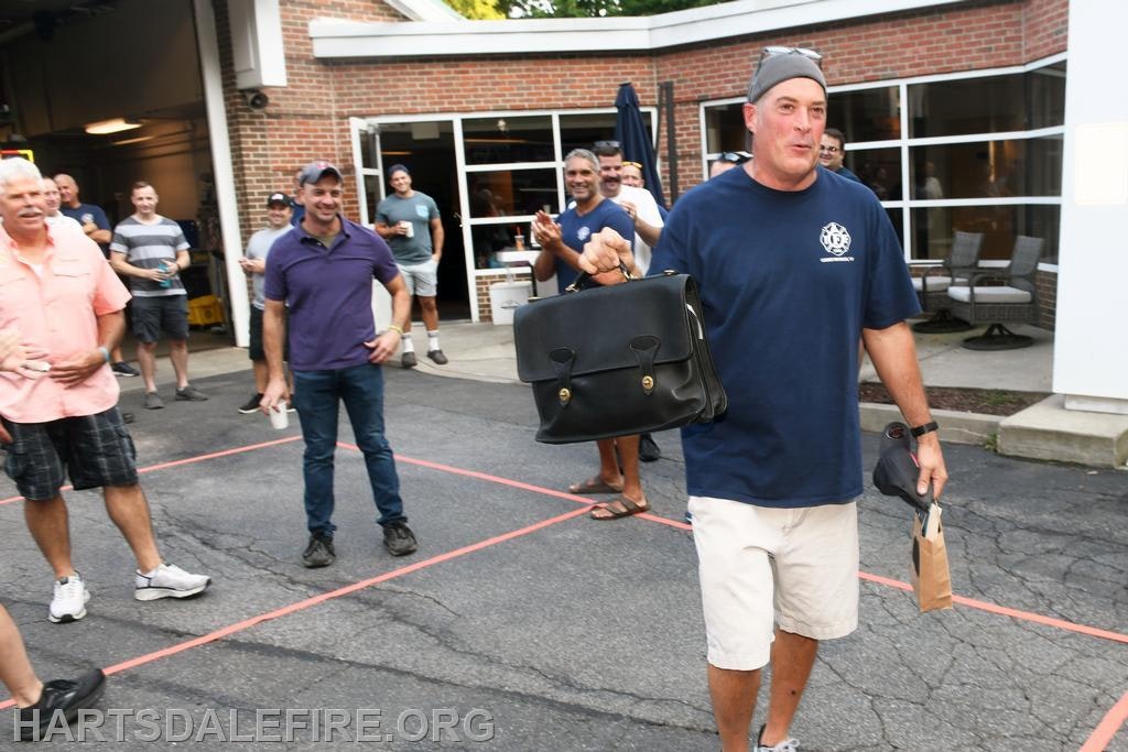 A group of men applauds as one man in a blue shirt and shorts walks with a bag, possibly celebrating an event.