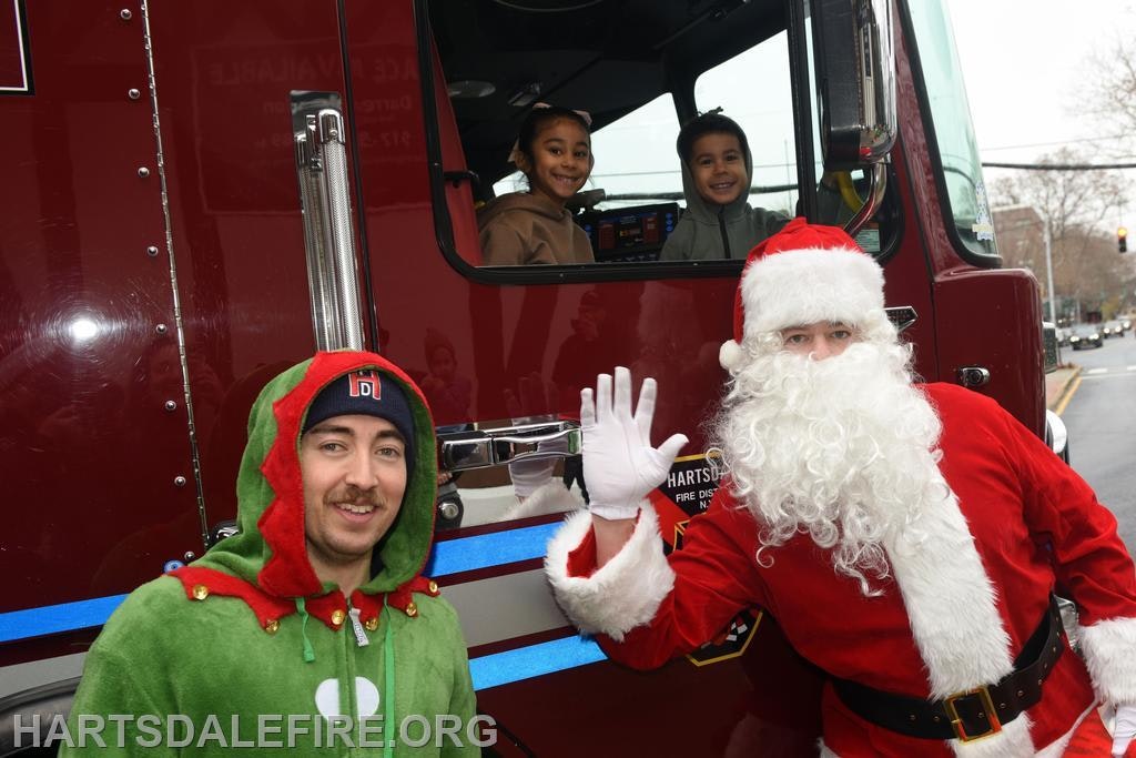 A man in an elf costume with Santa waves, while two children sit in a fire truck. It looks festive and cheerful!