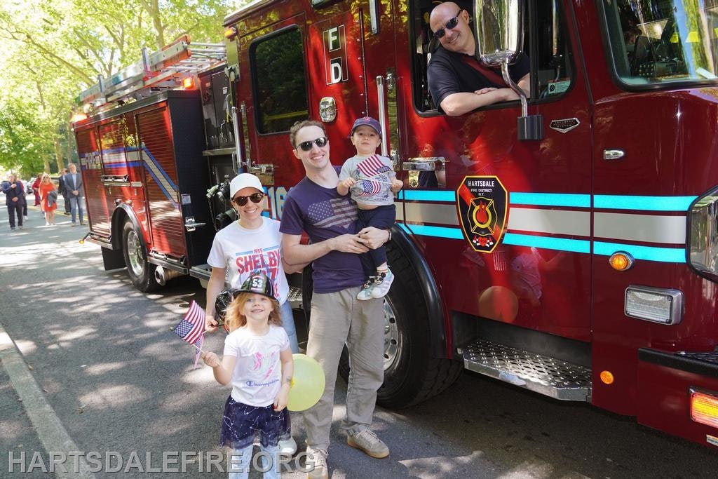 A family poses with children and a fire truck, celebrating a festive occasion, with flags and smiles all around.