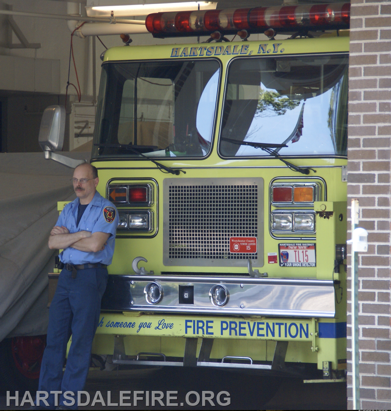 A fireman leans against a yellow fire truck in Hartsdale, NY, with a focus on fire prevention messaging.