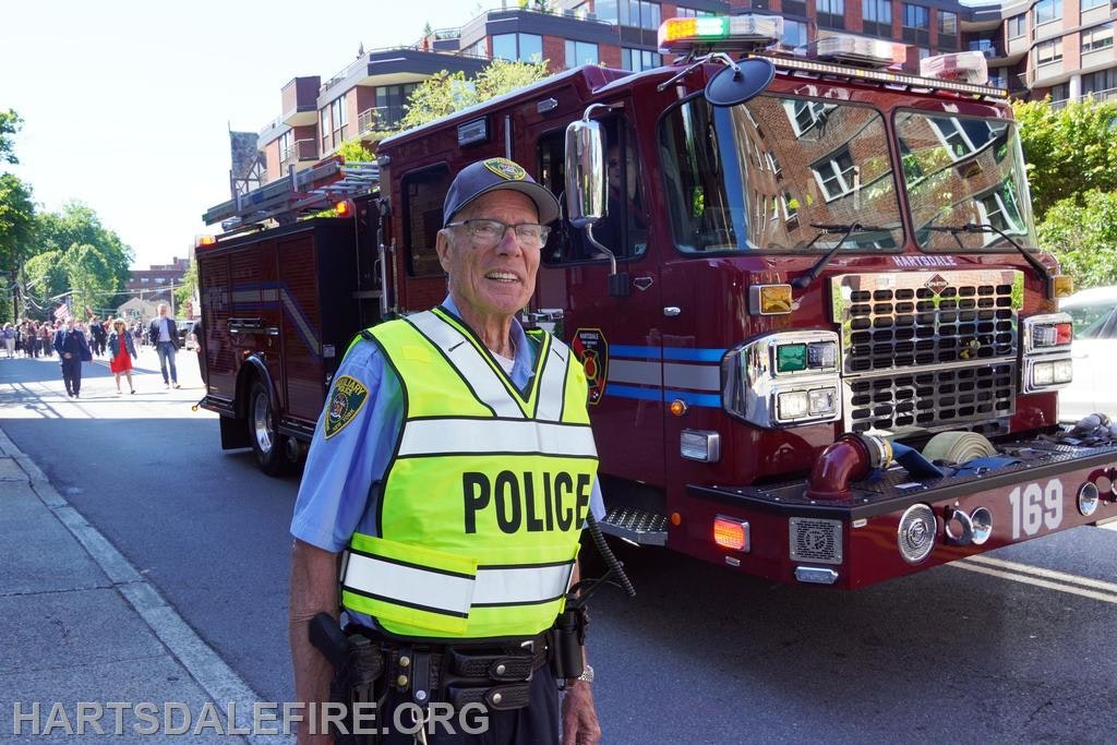 A police officer in a reflective vest stands in front of a fire truck, with a crowd walking in the background on a sunny day.