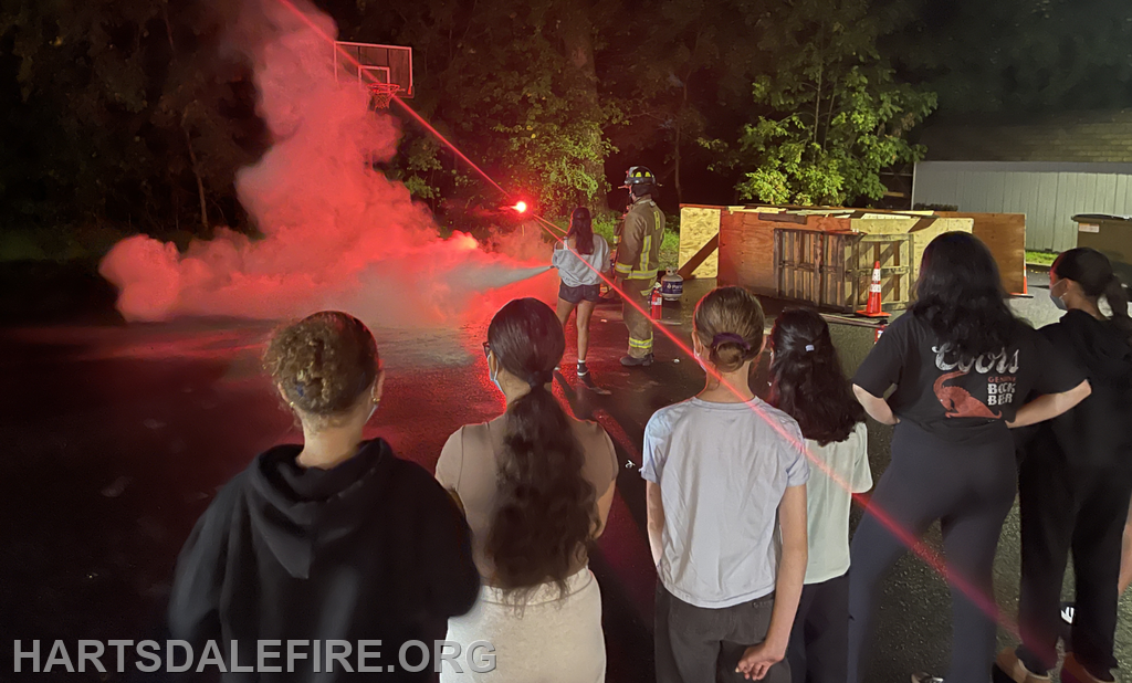 A group watches a firefighter demonstrate equipment in a smoky, night-time setting near a basketball hoop.