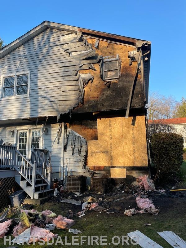 A house with significant fire damage on the exterior wall, with charred and melted siding. Debris is scattered on the ground.