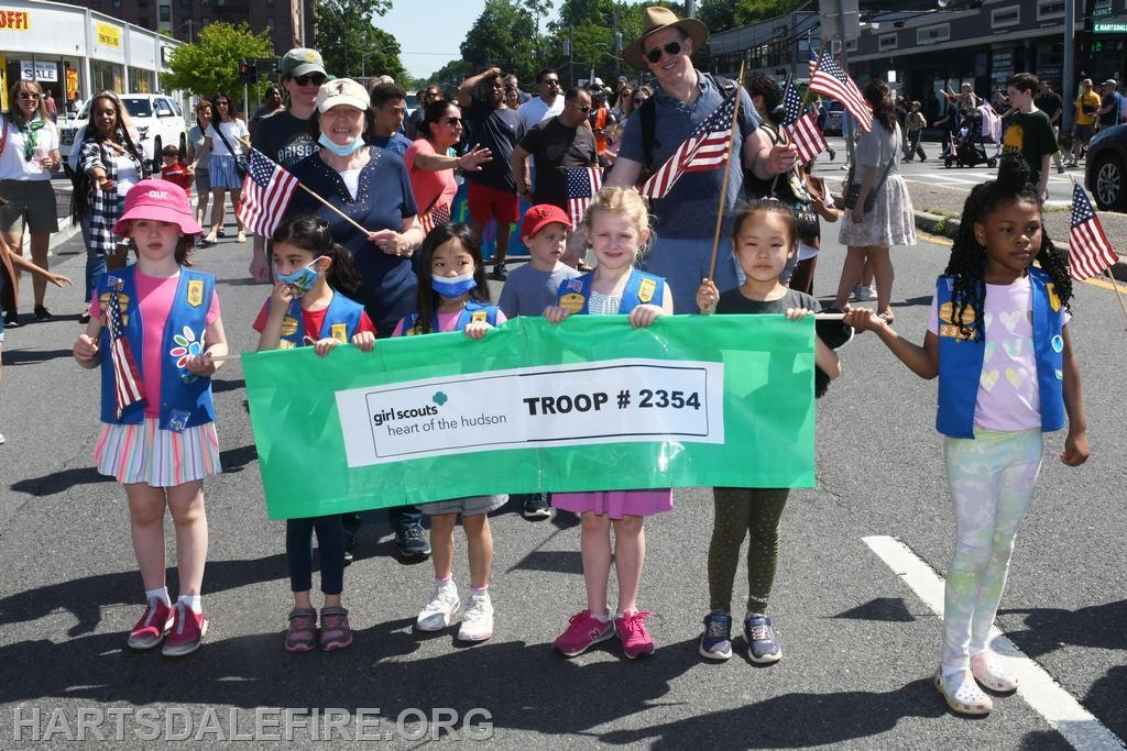 A group of children with a "Girl Scouts Troop #2354" banner in a parade, holding American flags.