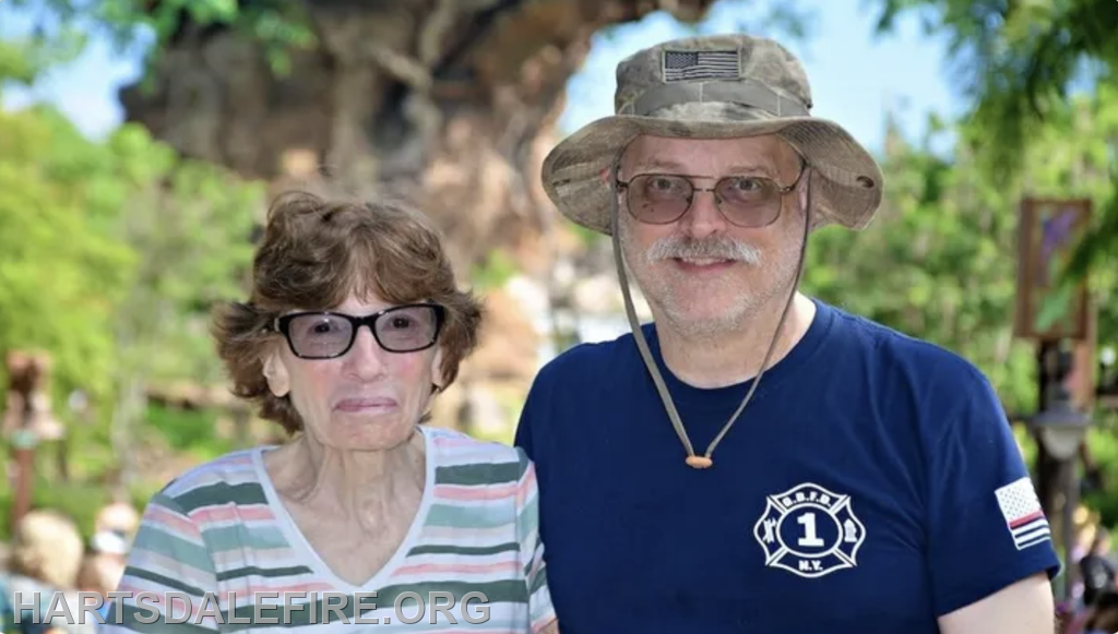A couple poses for a photo outdoors, smiling. The background features lush greenery, suggesting a sunny day.
