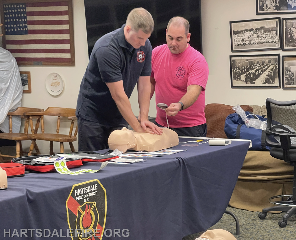 Two firefighters are practicing CPR on a training dummy in a classroom setting, with first aid materials on a table.